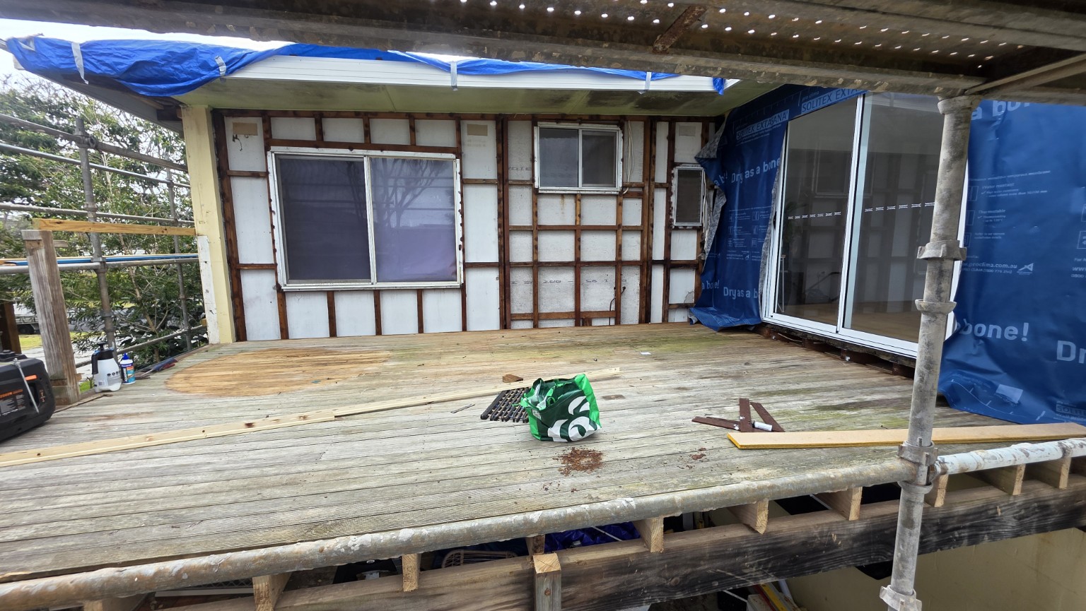 External wall cladding removed beside a timber deck, with existing windows retained and scaffold around the work area.